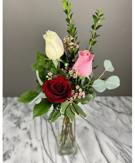 An arrangement of three roses in red, pink, and white with wax flowers, greenery, and eucalyptus stems, presented in a rectangular glass vase.