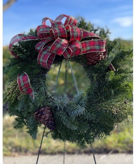 A fresh evergreen wreath decorated with pinecones and a red tartan plaid ribbon, designed for placement at a cemetery or memorial.