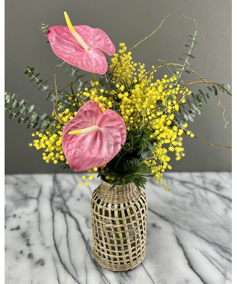 A spring floral arrangement featuring golden-yellow mimosa flowers, eucalyptus leaves, and willow branches in a natural rattan vase. Soft and rustic.